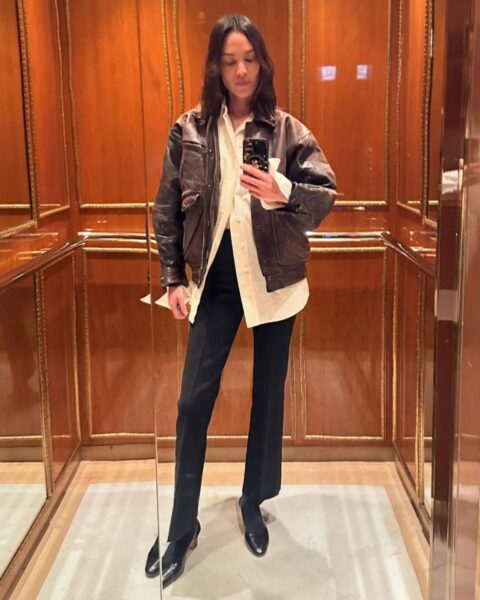 Zdjęcie modelki Alexy Chung, która zrobiła sobie selfie w windzie. Ma na sobie czarne spodnie dzwony, białą, luźną koszulę oraz kurtkę skórzaną. Na nogach czarne czółenka. Ma włosy ciemne, do ramion.