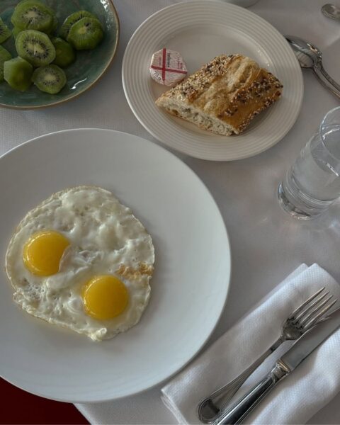 Zdjęcie stołu z białym obrusem, na którym stoją talerze z pieczywem, jajkami sadzonymi oraz pokrojonym kiwi. Te produkty to sposoby na gładszą szyję po 40.