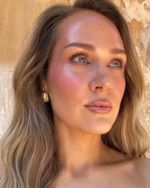Zdjęcie kobiety w długich, blond włosach rozpuszczonych na ramiona. Ma złote kolczyki w uszach. Na twarzy makijaż typu sunburnt blush. Patrzy w dal.