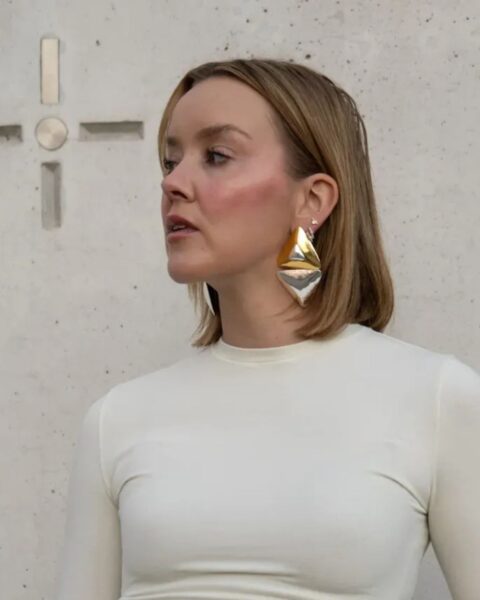 Zdjęcie kobiety z blond, prostymi włosami ściętych w stylu loba, czyli long boba. Ubrana jest w kremowy top z długim rękawem oraz białe jeansy. W uszach ma wiszące, złote kolczyki. Na ręce ma złotą bransoletkę, a na palcach pierścionki.
