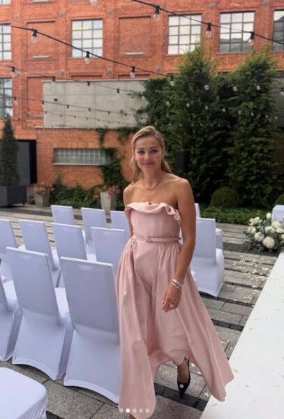 Kobieta z blond włosami w różowej sukience w klimacie new look. W tle białe krzesła i celglany budynek.