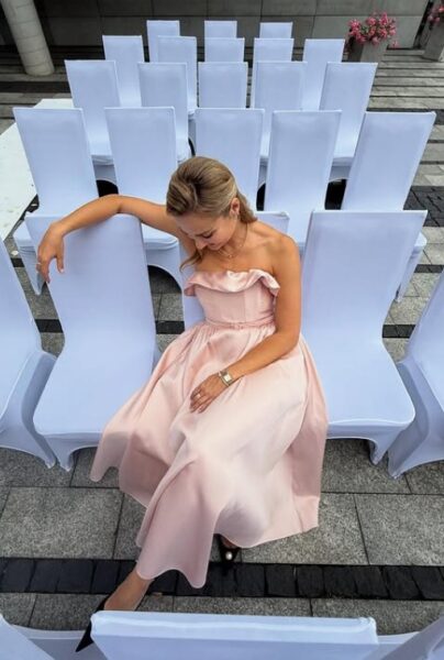 Kobieta z blond włosami i w różowej midi sukience w klimacie new look siedzi na białym krześle. Dookoła niej stoją inne, białe krzesła. Kobieta nie patrzy w obiektyw.