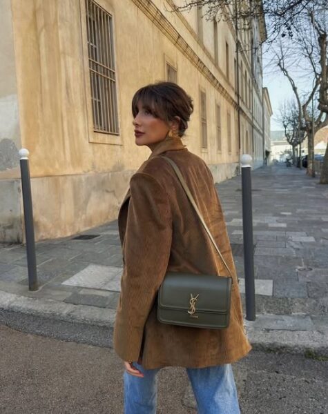 Kobieta w brązowej marynarce i niebieskich jeansach pozuje na tle miasta. Na ramieniu ma skórzaną torebkę.