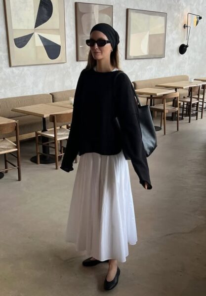 Kobieta w białej, długiej spódnicy i czarnym swetrze pozuje na tle pustej restauracji.