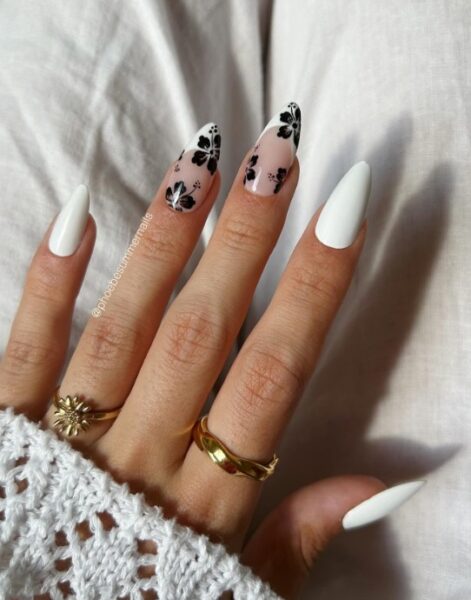 Biały manicure. Dwa paznokcie pomalowane na jasny odcień. Na tych płytkach znajdują się czarne kwiaty - hibiskusy.