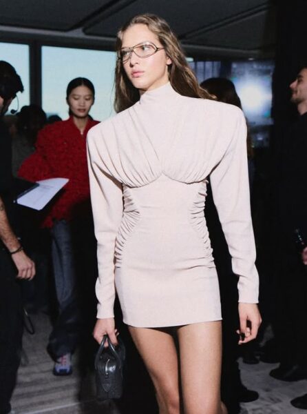 Modelka na backstage w jasnej sukience z poszerzanymi ramionami.