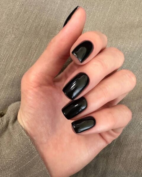 Zdjęcie dłoni ze zbliżeniem na czarny manicure, który należy do 10 najpopularniejszych kolorów paznokci wszech czasów. Paznokcie są średniej długości, kwadratowe. Dłoń pozuje na tle brązowej marynarki.