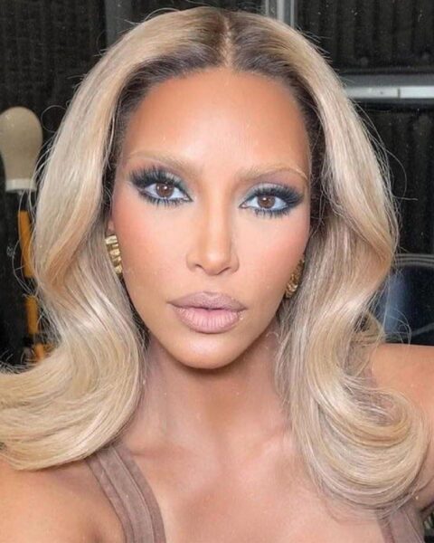 Zdjęcie celebrytki Kim Kardashian w blond włosach z podkręconymi końcówkami i jasnymi brwiami oraz mocnym makijażem twarzy. Ubrana jest w beżowy top. W uszach złote kolczyki.