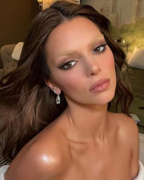 Zdjęcie celebrytki Kendall Jenner w trakcie przygotowań do Met Gala. Jest ubrana w biały szlafrok, ma ciemne, falowane, rozpuszczone na plecy włosy. Ma mocny makijaż twarzy, szczególnie oczy oraz jasne brwi. W uszach srebrne kolczyki.