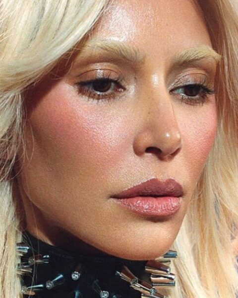 Zdjęcie portretowe Kim Kardashian, która na zdjęciu ma blond włosy z grzywką na boki oraz rozjaśniane brwi. Ma na szyi obrożę z kolcami.