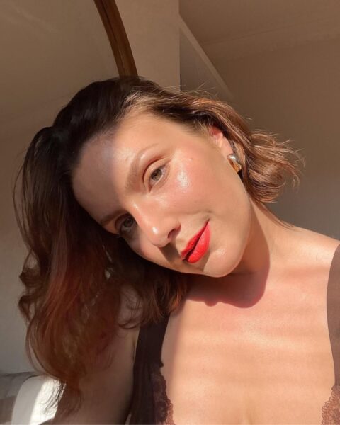 Zdjęcie kobiety, która zrobiła sobie selfie. Przechyla głowe na bok. Ma ciemne, krótkie włosy do ramion. Jest ubrana w brązowy top na ramiączkach z koronką. Ma makijaż z czerwonymi ustami, który zmyje wieczorem, bo wie, co się stanie gdy nie zmyjesz makijażu. Pozuje przy ścianie, obok lustra.