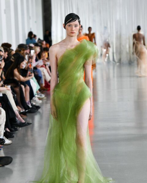 Zdjęcie z pokazu haute couture PFW - Anatomy Of Identity By Lever Runway. Na wybiegu idzie modelka w zielonej kreacji z rozcięciem i jednym ramiączkiem. Suknia ma nietypową formę i powstała z tiulu. Ma ciemne włosy gładko zaczesane.