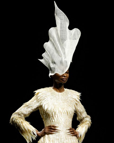 Zdjęcie z pokazu haute couture Robert Wun Couture AW25. Ciemnoskóra modelka jest ubrana w kremową suknię z piór, a na głowie ma unikalny kapelusz w kształcie muszli.