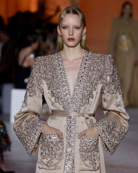 Zdjęcie z pokazu haute couture Zuhair Murad Couture AW25. Blond modelka ubrana jest w kremowy płaszcz z paskiem w talii i kieszeniami. Całość jest wyszywana kryształami. Modelka ma blond włosy rozpuszczone na plecy.