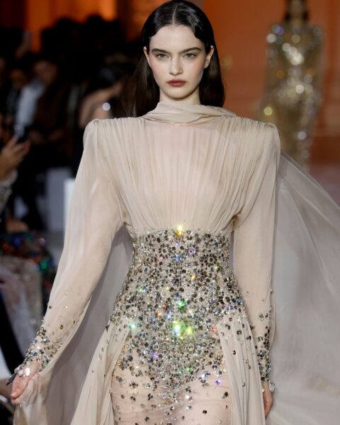 Zdjęcie z pokazu haute couture Zuhair Murad Couture AW25. Modelka ma ciemne włosy rozpuszczone na ramiona. Ubrana jest w kremowa sukienkę plisowaną z tiulu i z talią wyszywaną kamieniami.