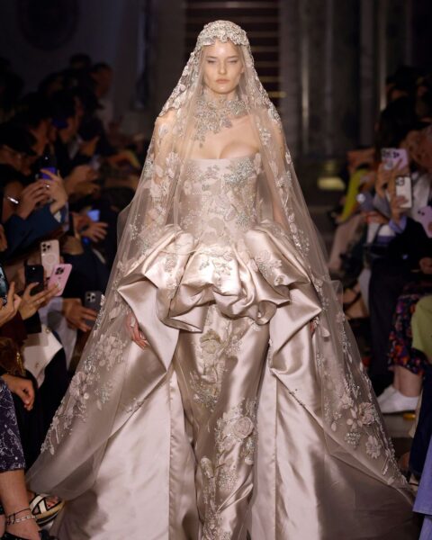 Zdjęcie z pokazu haute couture Elie Saab Couture AW25. Modelka ma na sobie złotą suknię wieczorową z baskinką i gorsetem oraz szerokim trenem. Ma na głowie długi welon z koronką.