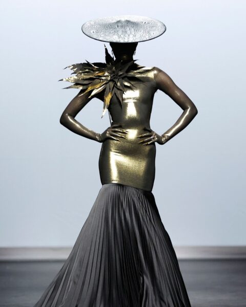 Zdjęcie z pokazu haute couture Robert Wun Couture AW25. Kobieta jest ubrana w suknię typu syrena w kolorze złotym, plisowanym, czarnym dołem oraz kwiatem na ramieniu. Do tego kobieta ma płaski kapelusz na głowie.