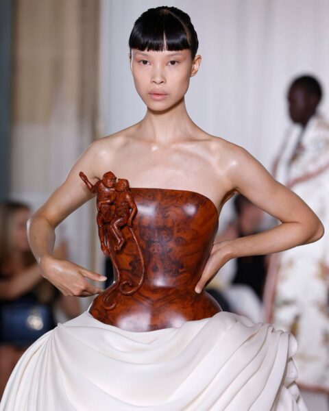 Zdjęcie z pokazu haute couture Ashi Couture AW25. Modelka ma ciemne włosy związane gładko i prostą grzywkę. Ubrana jest w kreację z gorsetem z drewna i charakterystycznym zdobieniem oraz białą spódnicą.