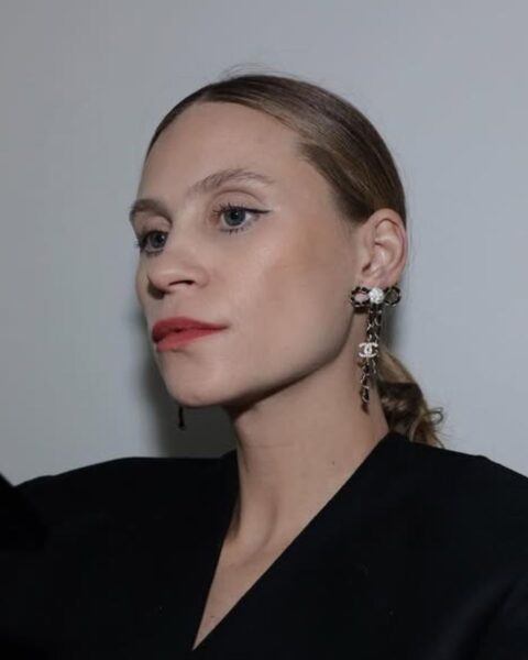 Zdjęcie kobiety z blond włosami, gładko związanymi z tyłu. Jest ubrana w czarną bluzkę. W uszach ma wiszące, długie, lśniące kolczyki. Patrzy w bok.
