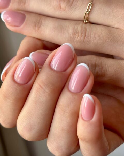 Zdjęcie dłoni ze zbliżeniem na krótkie, owalne paznokcie z soft french manicure w kolorze różowym i średniej grubości białą końcówką. Widać na palcu pierścionek w kolorze złotym. Dłonie układają się tak, by prezentować manicure.