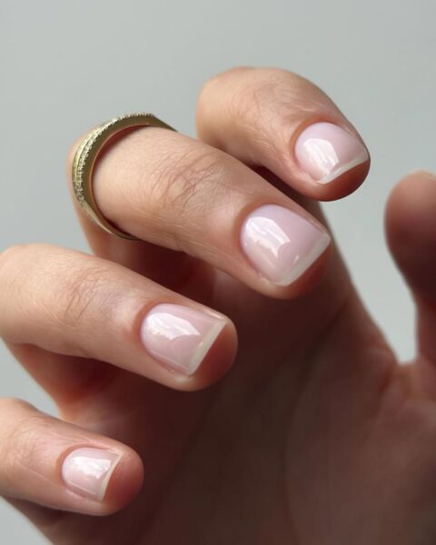 Zdjęcie dłoni ze zbliżeniem na krótkie paznokcie z french manicure z efektem soap nails, czyli mydlane paznokcie. Na palcu środkowym widać złoty pierścionek.
