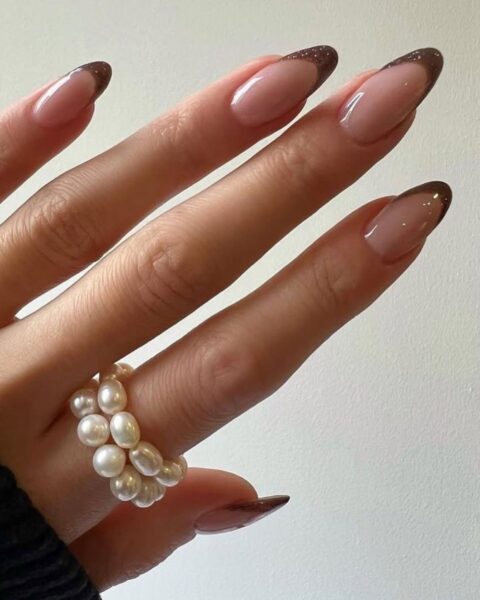 Zdjęcie dłoni ze zbliżeniem na długie, migdałowe paznokcie z french manicure z brązową końcówką. Na palcu wskazującym widać podwójny pierścionek z perełek.