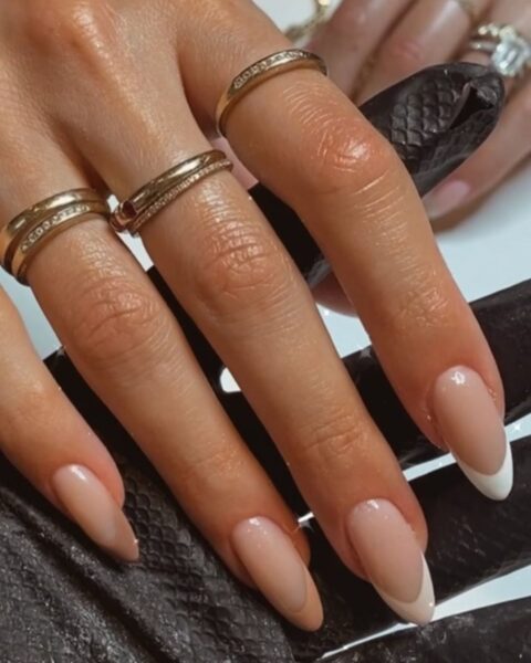 Zdjęcie dłoni ze zbliżeniem na długie, migdałowe paznokcie. Są pomalowane w stylu french manicure z beżowymi końcówkami, zamiast białych. Na każdym palcu widać złoty pierścionek.