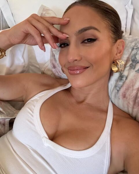 Zdjęcie Jennifer Lopez leżącej na kanapie. Jest ubrana w biały top, przykłada rękę do twarzy i widać jej french manicure. Ma w uszach złote pierścionki. Włosy związane gładko. Szeroko się uśmiecha.