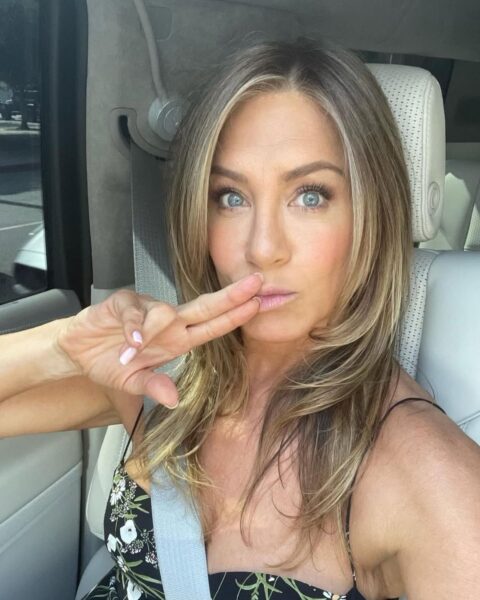 Zdjęcie Jennifer Aniston, która zrobiła sobie selfie w samochodzie. Ma rozpuszczone blond włosy. Ubrana jest w czarą sukienkę w kwiatki na ramiączkach. Przykłada rękę do twarzy. Widać jej mleczy french manicure.