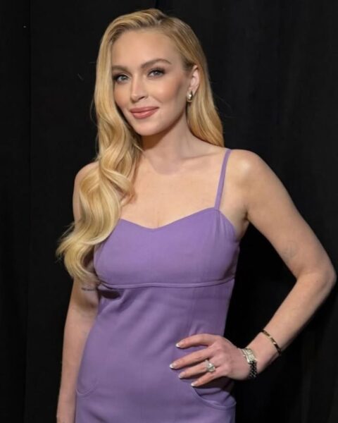 Zdjęcie Lindsay Lohan. Jest ubrana w fioletowa mini sukienkę na ramiączkach. Ma długie, rozpuszczone blond włosy. Na rękach bransoletki i pierścionki. Nosi klasyczny french manicure. Pozuje na czarnym tle.