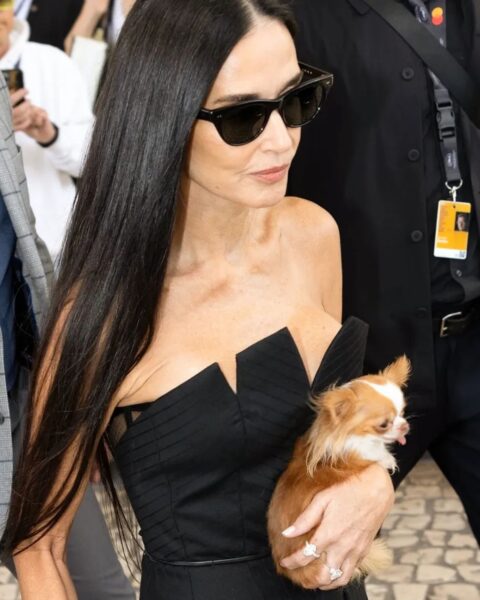 Zdjęcie Demi Moore. Ma ciemne, rozpuszczone włosy. Okulary przeciwsłoneczne na nosie. Jest ubrana w czarną, wieczorową suknię bez ramiączek. W rękach trzyma pieska. Widać jej french manicure.