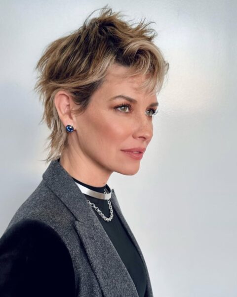 Zdjęcie kobiety w niemodnej na jesień 2025 fryzurze typu pixie cut, mocno cieniowanej. Ubrana jest w czarną bluzkę i szarą marynarkę. Ma srebrny, podwójny naszyjnik.