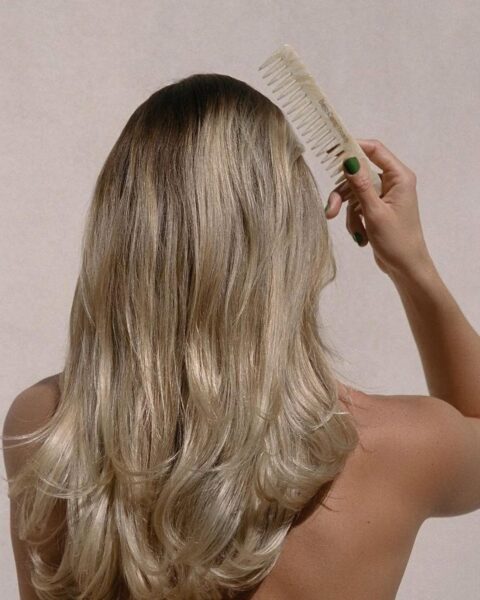 Zdjęcie kobiety stojącej tyłem. Czesze swoje długie, blond włosy po zabiegu glass hair. Czesze się beżowym grzebieniem.