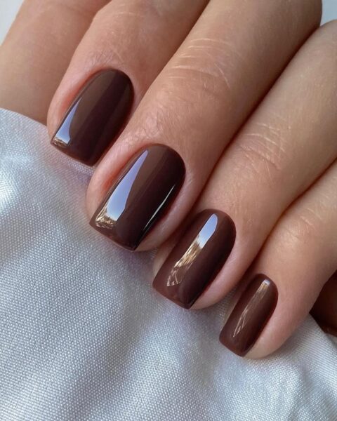Zdjęcie ze zbliżeniem na długie, kwadratowe paznokcie. Są pomalowane lakierem w kolorze czekoladowego brązu. Dłoń pozuje tak, by prezentować manicure.