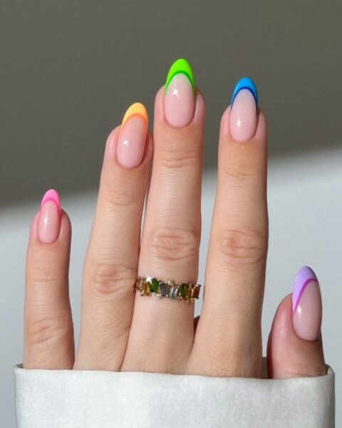 Zdjęcie dłoni z długimi, migdałowymi paznokciami z podwójnym french manicure z kolorowymi końcówkami w stylu jelly. Na palcu środkowym widać złotą obrączkę z zielonymi kamieniami.