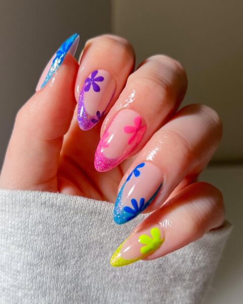 Zdjęcie dłoni z długimi, migdałowymi paznokciami z kolorowym, żellkowym french manicure z brokatem oraz zdobieniem kwiatów.