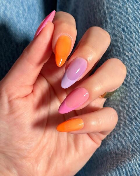 Zdjęcie dłoni ze zbliżeniem na długie, migdałowe paznokcie w stylu french manicure z żelkowym efektem i neonową końcówką. Paznokcie mają kolor pomarańczu, fioletu i różu.