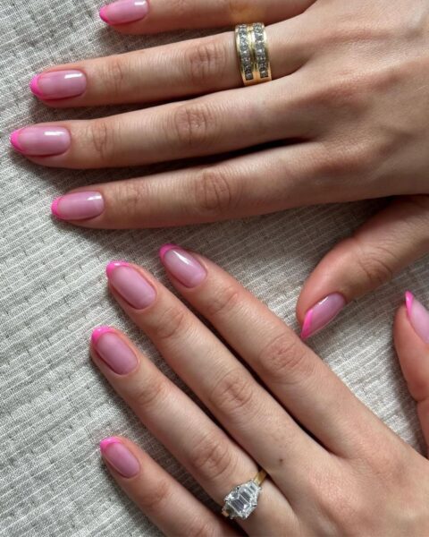 Zdjęcie dłoni z długimi, owalnymi paznokciami pomalowanymi w stylu french manicure. Na palcach widać masywną, złotą obrączkę i srebrny pierścionek z brylantem. Dłonie opierają się o beżowy kocyk.