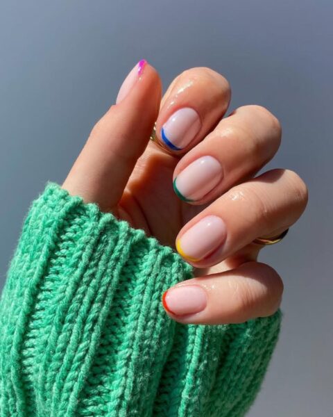 Zdjęcie dłoni z krótkimi, owalnymi paznokciami z żelkowym micro french manicure, gdzie każda końcówka ma inny kolor. Widać fragment rękawa zielonego swetra.