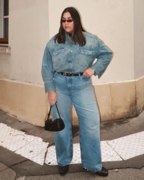 Zdjęcie kobiety plus size ubranej w jesienne trendy, które wyszczuplają: jeansowe spodnie z wysokim stanem i szeroką, jeansową koszulę. Ma w pasie pasek, w ręce czarną małą torebkę, na nogach czarne buty. Włosy ciemne, rozpuszczone, do ramion. Na nosie ma okulary przeciwsłoneczne. Stoi na ulicy.