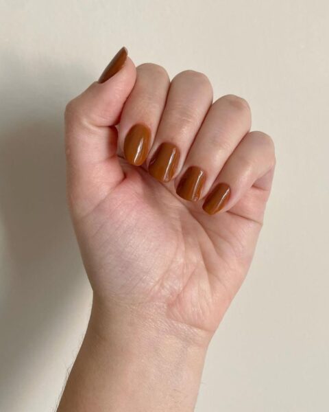Zdjęcie dłoni ze zbliżeniem na średniej długości, kwadratowe paznokcie, które zdobi karmelowy manicure. Dłoń pozuje na tle białej ściany.