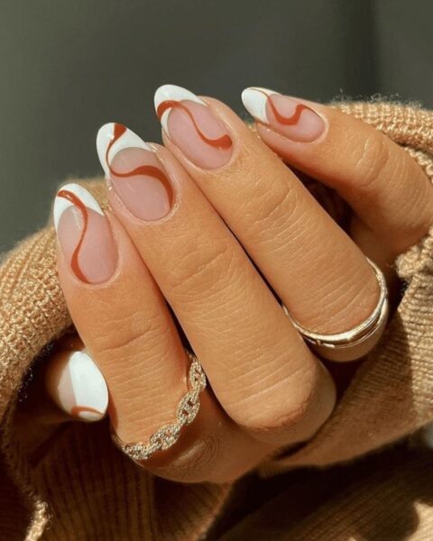 Zdjęcie dłoni z długimi, owalnymi paznokciami z french manicure i wzorkiem swirl w kolorze karmelowego brązu. Na palcach widać złote pierścionki.