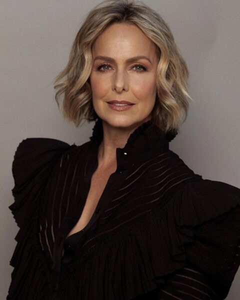Zdjęcie aktorki Melory Hardin. Ma krótkie włosy typu bob, falowane. Pozuje do zdjęcia ubrana w czarną marynarkę z szerokimi ramionami z poduszkami.