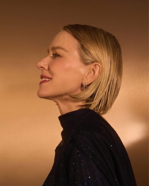 Zdjęcie aktorki Naomi Watts pozującej od boku. Jest ubrana w czarną sukienkę ze stójką, a jej materiał jest wyszywany cekinami. Ma krótkie blond włosy w stylu sleek boba.