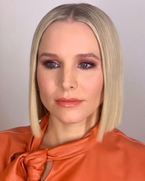 Zdjęcie aktorki Kristen Bell. Ma włosy typu blunt bob w kolorze blond. Jest ubrana w pomarańczową bluzkę z wiązaniem przy szyi.
