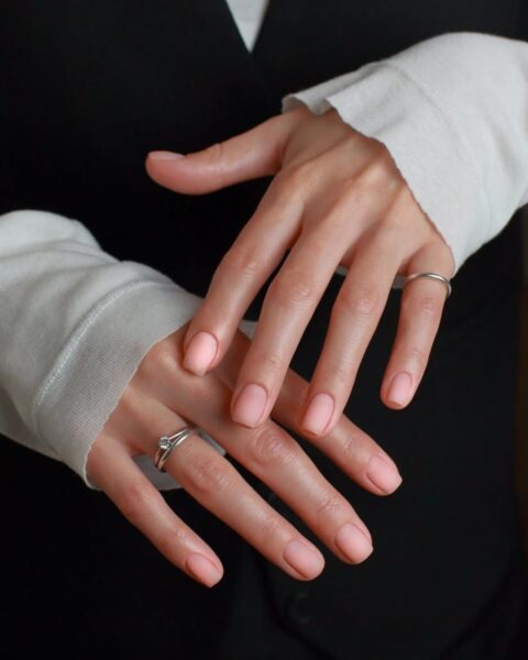 Zdjęcie przedstawiające manicure, który odwraca uwagę od pomarszczonych dłoni. Widać kobietę wystawiającą dłonie do przodu, mają paznokcie pomalowane na kolor nude. Na palcu serdecznym jednej z dłoni znajduje się srebrny pierścionek. Widać, że kobieta ma na sobie białą bluzkę oraz czarną kamizelkę.