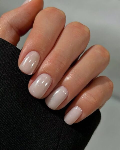 Zdjęcie dłoni ze zbliżeniem na odmładzający manicure. Paznokcie są średniej długości, mają kształt zaokrąglonego kwadratu i są pomalowane na kolor biały z drobinkami. Widać fragment rękawa czarnej bluzki.