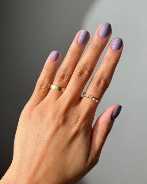Zdjęcie ze zbliżeniem na manicure, który odmładza: średniej długości kwadratowe paznokcie w kolorze brudnego fioletu. Na palcach widać dwa złote pierścionki.