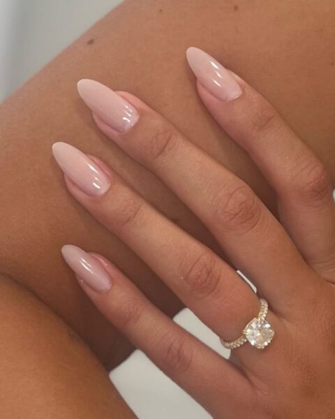 Zdjęcie ze zbliżeniem na manicure, który postarza: długie, migdałowe paznokcie w kolorze nude. Na palcu widać złoty pierścionek z dużym brylantem.
