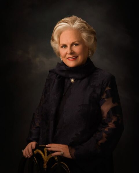 Zdjęcie Jacqueline Mars. Pozuje do portretu. Ma krótkie, blond włosy. ubrana jest w koronkową, czarną bluzkę, na szyi ma czarny szal i przypiętą przy nim broszkę. Opiera się dłońmi o bogato zdobione krzesło.